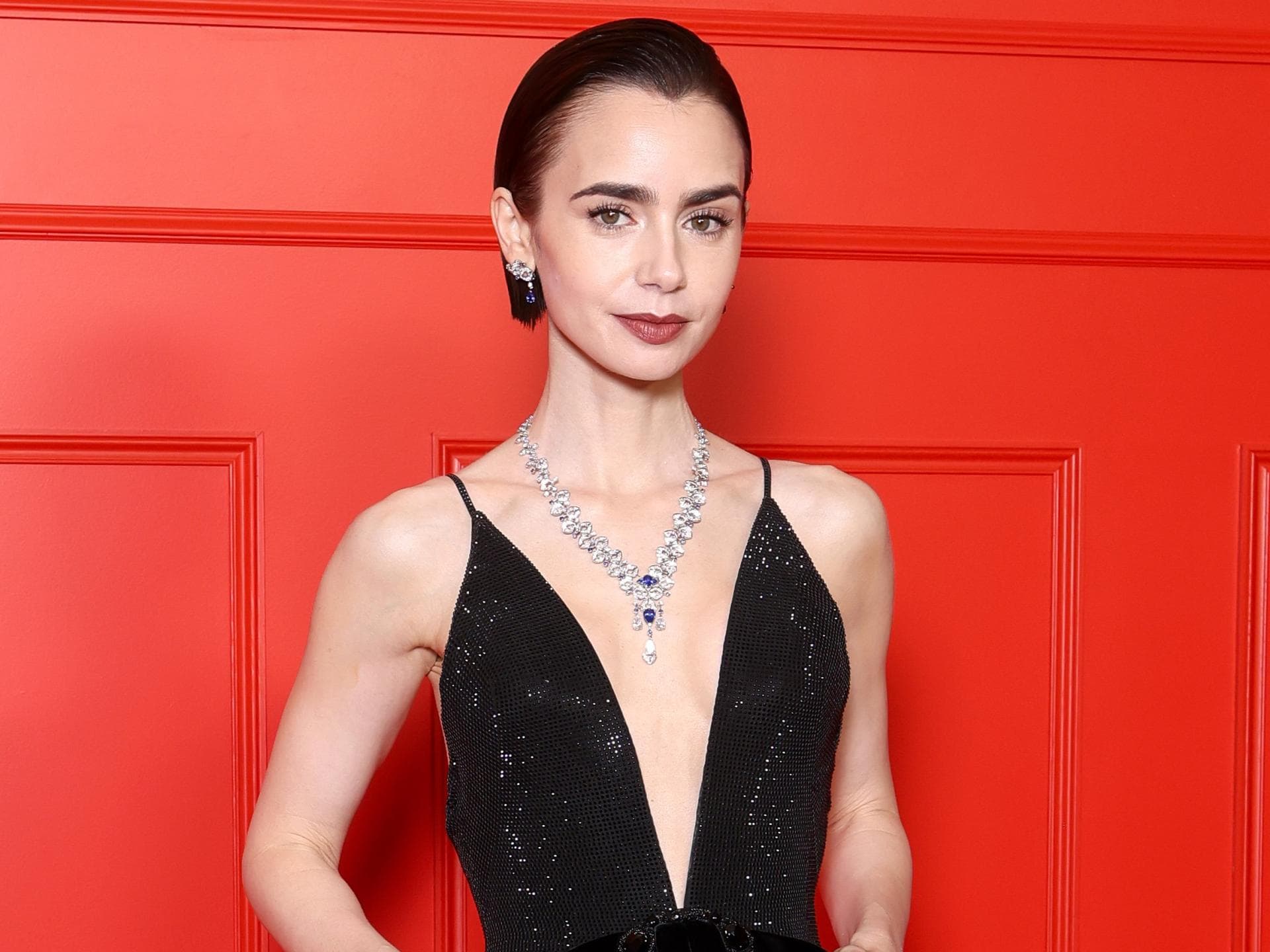 Lily Collins wird optisch oft mit Audrey Hepburn in Verbindung gebracht. Lily Collins in einem schwarzen Abendkleid mit tiefem V-Ausschnitt vor einer roten Wand.
