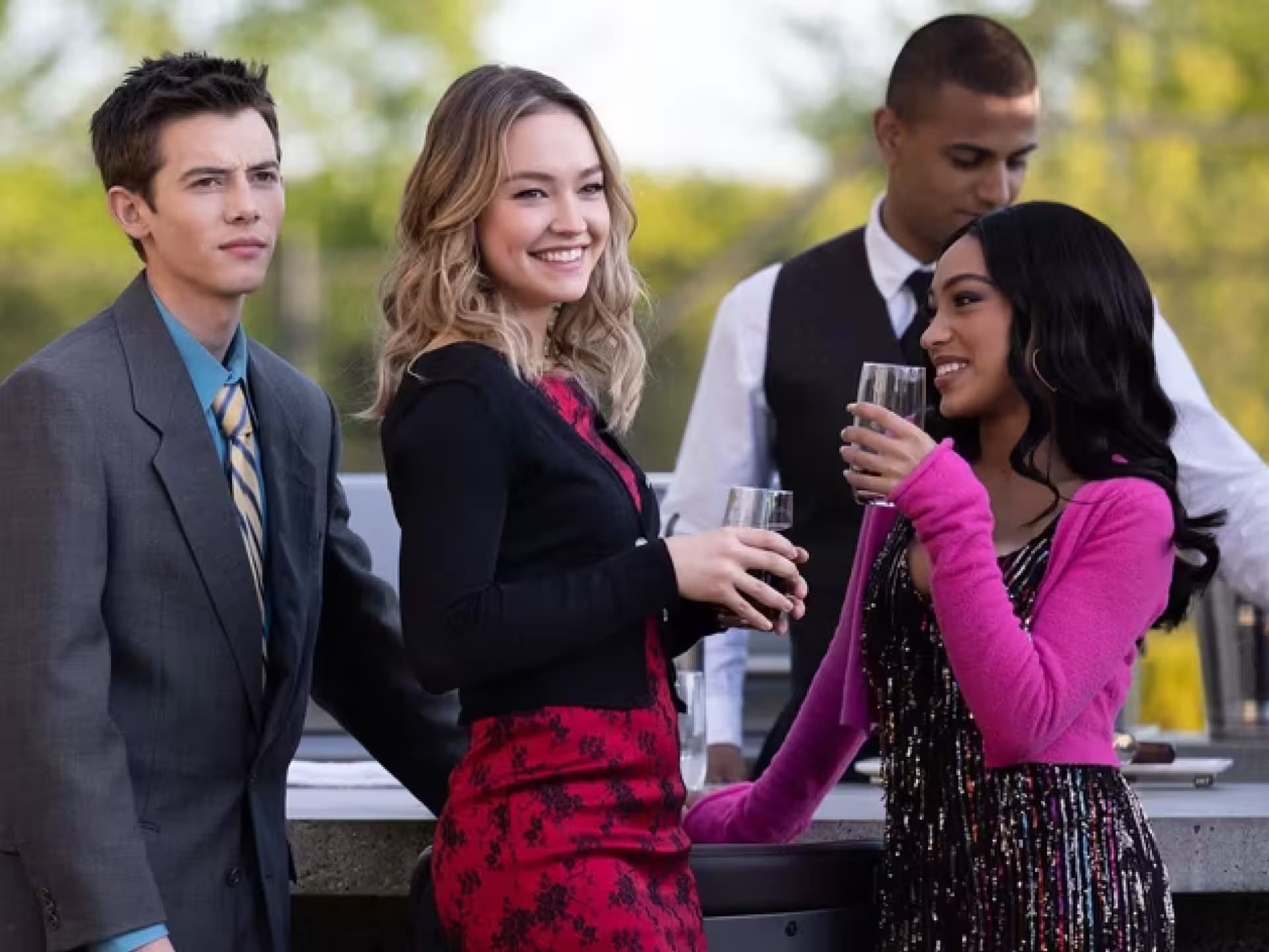 Drei Teenager in schicker Kleidung stehen draußen an einem Getränkestand, an dem ein Kellner arbeitet.