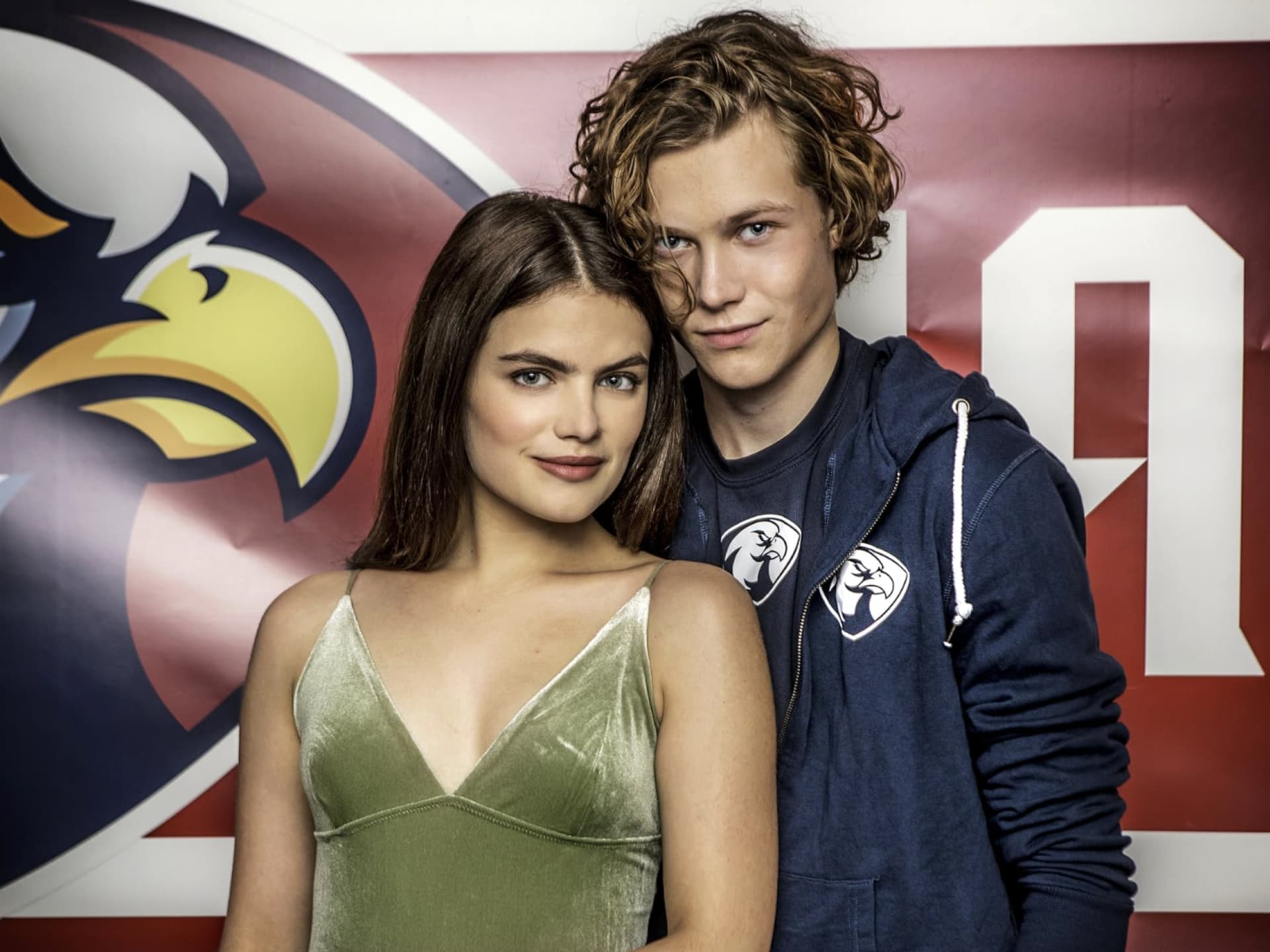 Eine junge Frau und ein junger Mann stehen vor einer Wand mit einem Adler-Banner.