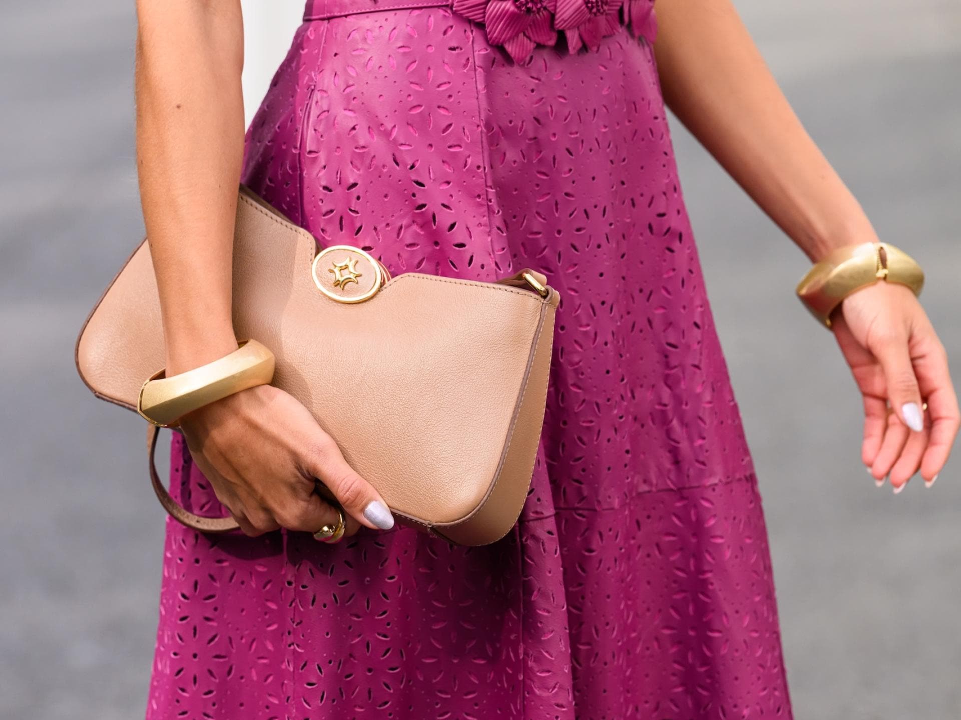 Eine Nahaufnahme einer Frau mit einem Kleid mit Lochmuster in Magenta und einer hellbraunen Clutch in der Hand mit goldenen Armreifen.