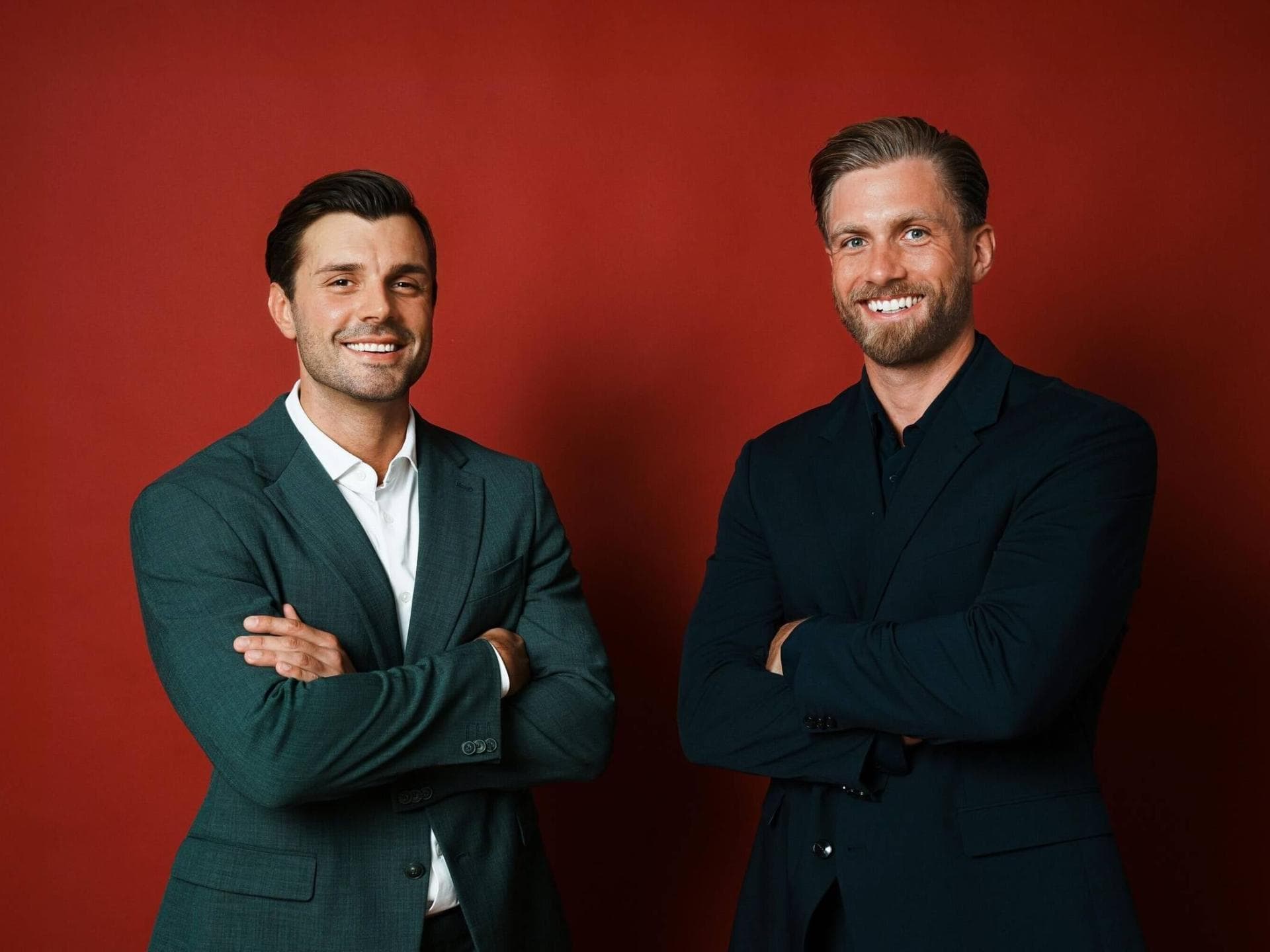 Tim Reitz und Sebastian Paul in Anzügen mit verschränkten Armen vor einer roten Wand.