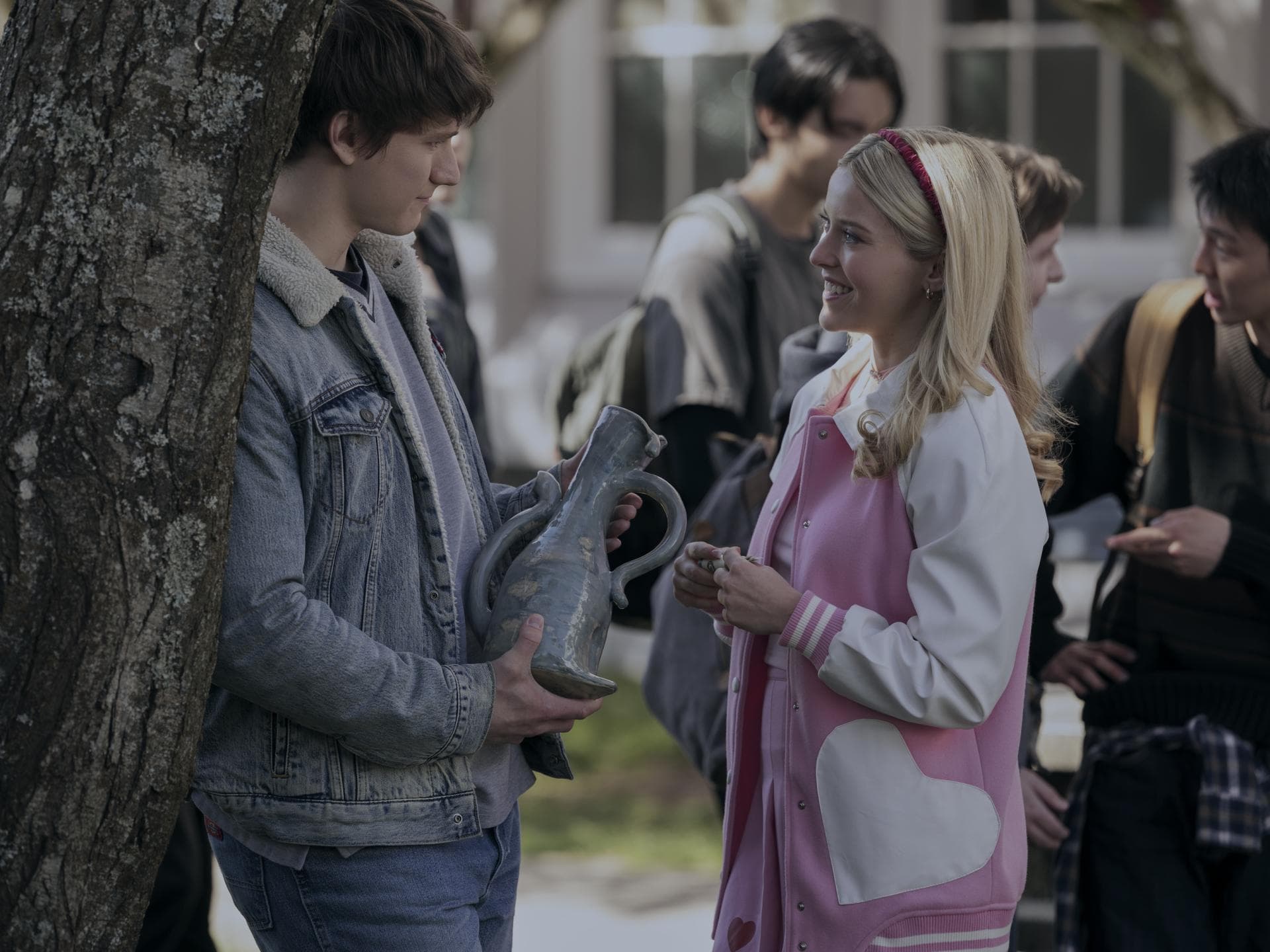 Eine junge Frau mit einer pink-weißen Bomberjacke und blonden Haaren spricht draußen mit einem jungen Mann in einer Jeansjacke, der eine Vase in der Hand hält.