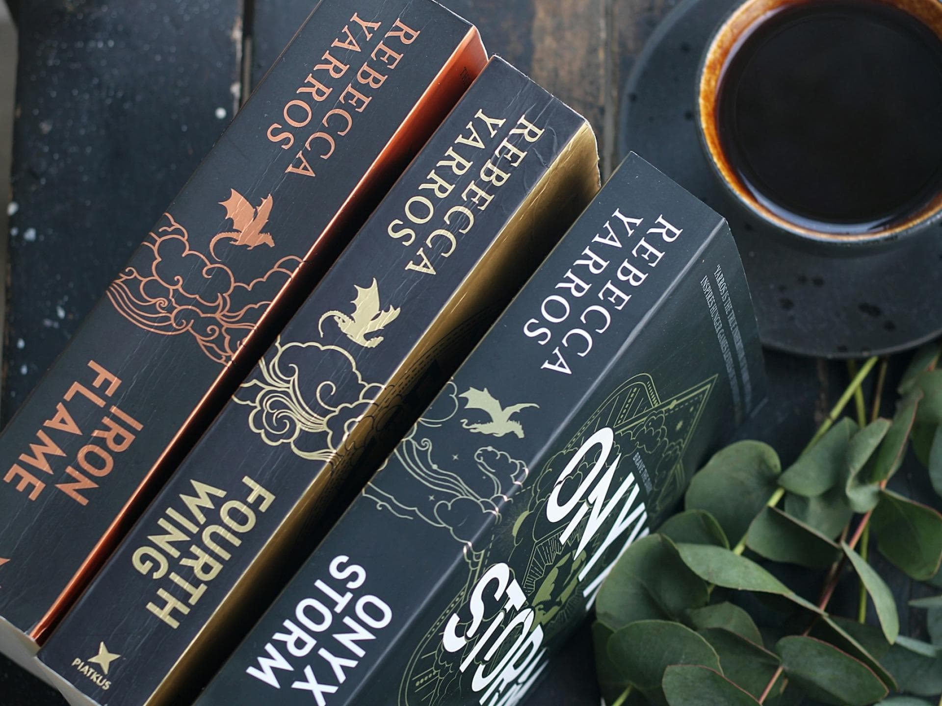 Drei Bücher liegen hochkant auf einem Tisch mit einer Tasse Kaffee und Eukalyptus-Sträuchern.