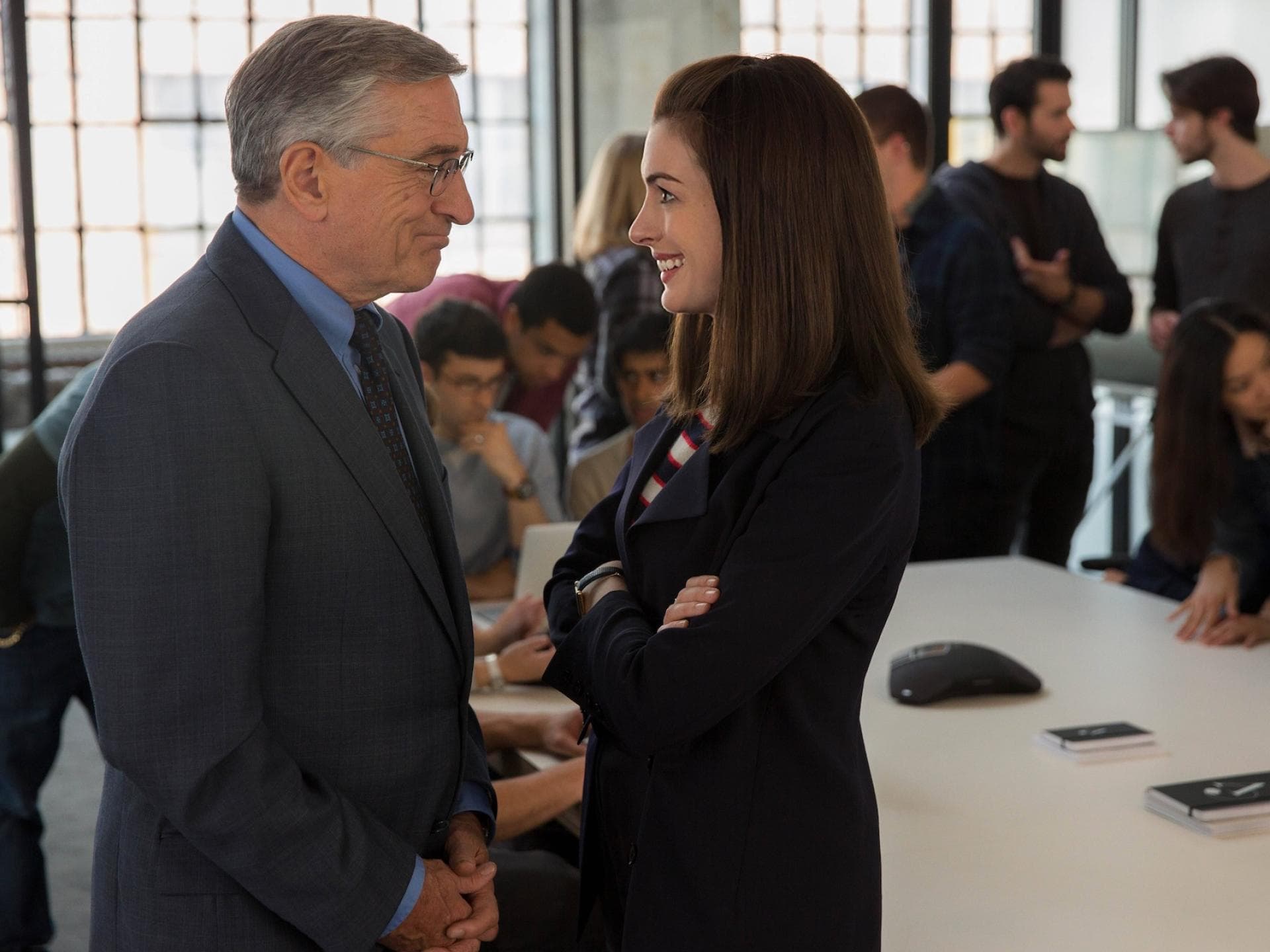 Anne Hathaway begeistert auch in „The Intern“ die Fans – hier an der Seite von Robert de Niro. Anne Hathaway und Robert de Niro stehen in einem vollen Büro und lächeln sich an.