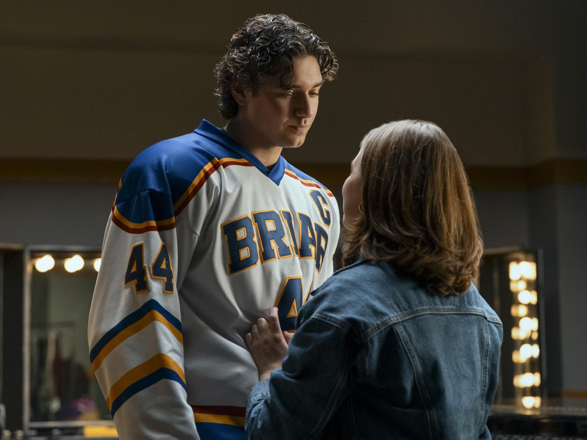 Ein junger Mann im Eishockey-Trikot steht einer jungen Frau in Jeansjacke gegenüber und guckt zu ihr herunter.