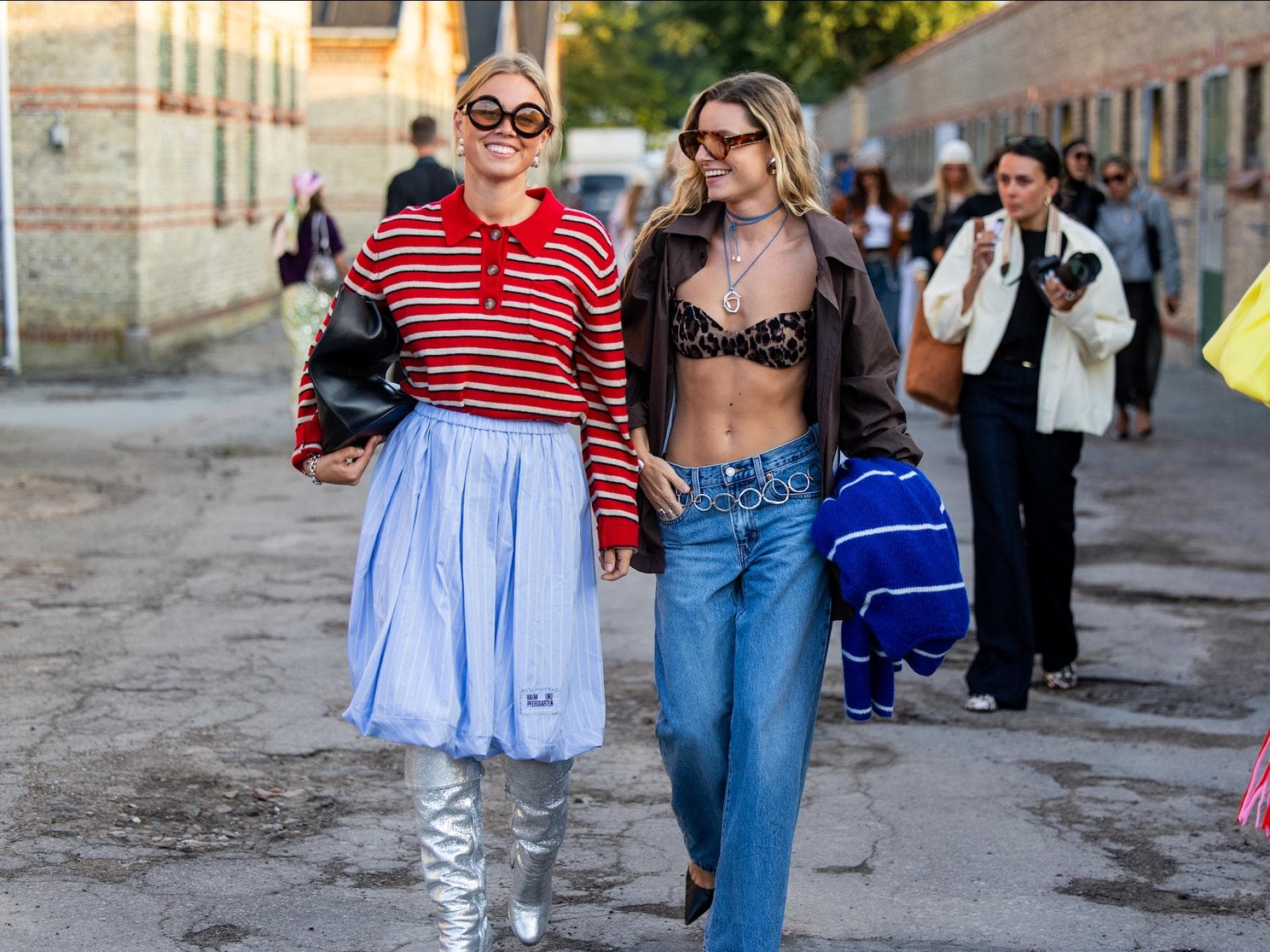 Zwei Frauen am Schlendern in der Stadt, eine mit einem Polo-Langarmshirt und einem blauen Rock, die andere mit einer Jeans und einem Leo-Bikini-Oberteil.