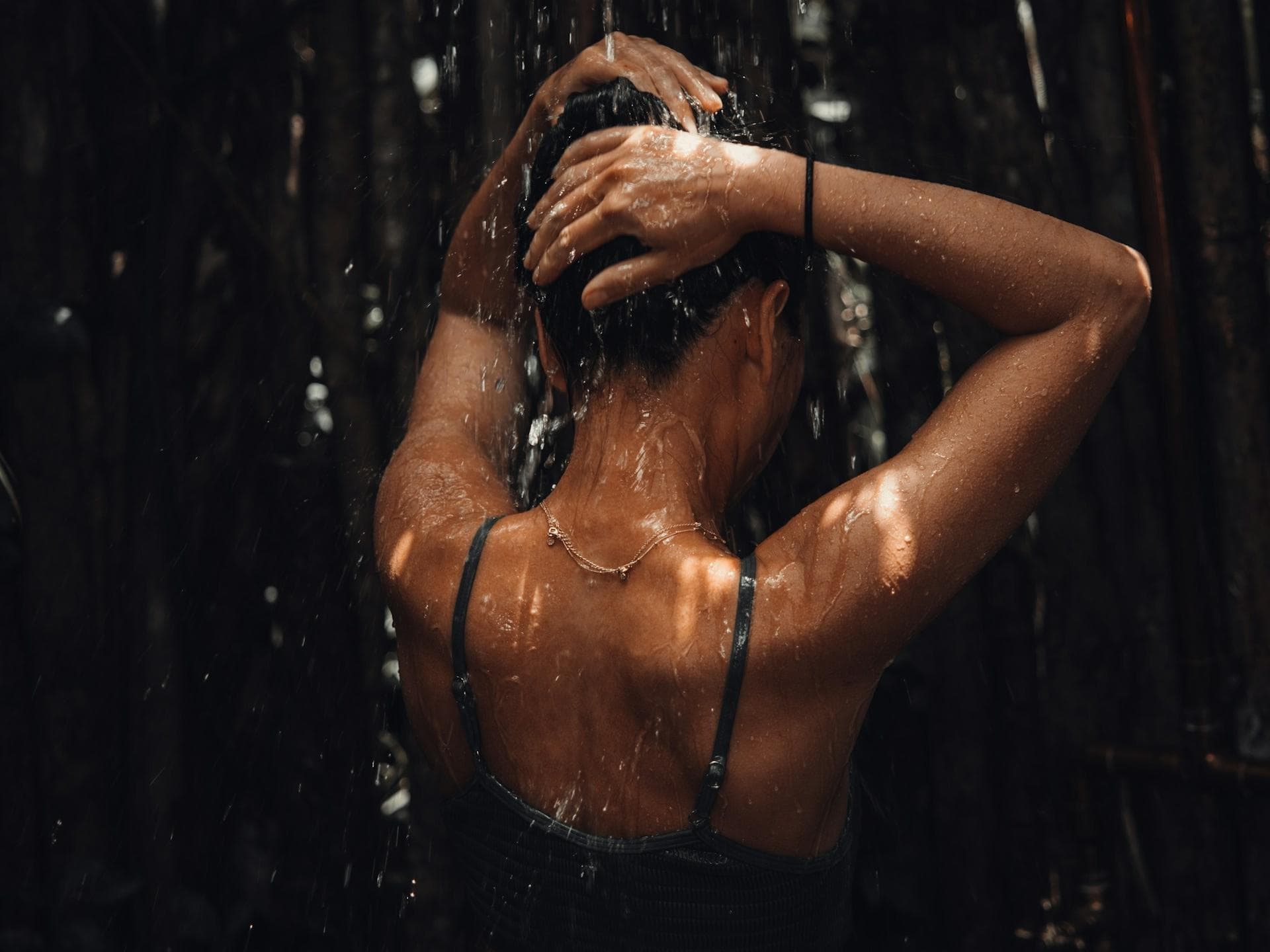Dark Showering soll den Körper optimal auf den Schlaf vorbereiten. Eine Frau steht mit dem Rücken zur Kamera in einer Dusche und lässt sich mit den Armen über dem Kopf berieseln.