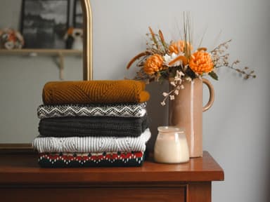 Gefaltete Wollpullover auf einer Holkommode neben einer Kerze und einer Blumenvase