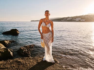 Eine junge Frau steht auf einem Felsen im Wasser und trägt einen weißen Bikini und einen Spitzenrock darüber.