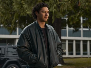 Ein junger Mann mit dunklen Locken in einer Lederjacke steht mit seinen Händen in der Tasche und guckt zur Seite.