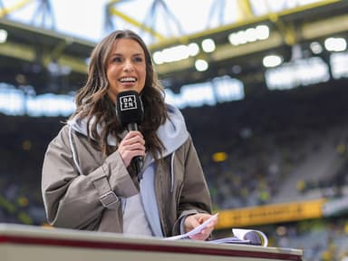 Eine junge Frau mit braunen Haaren steht in einem Fußballstadio und moderiert.