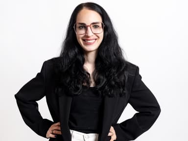 Michelle Calios ist Gründerin, Supply Chain Expertin und Business Angel. Eine Frau mit schwarzen Haaren, einem schwarzen Blazer und weißer Hose sowie einer Brille mit auf die Hüfte gestemmten Armen.